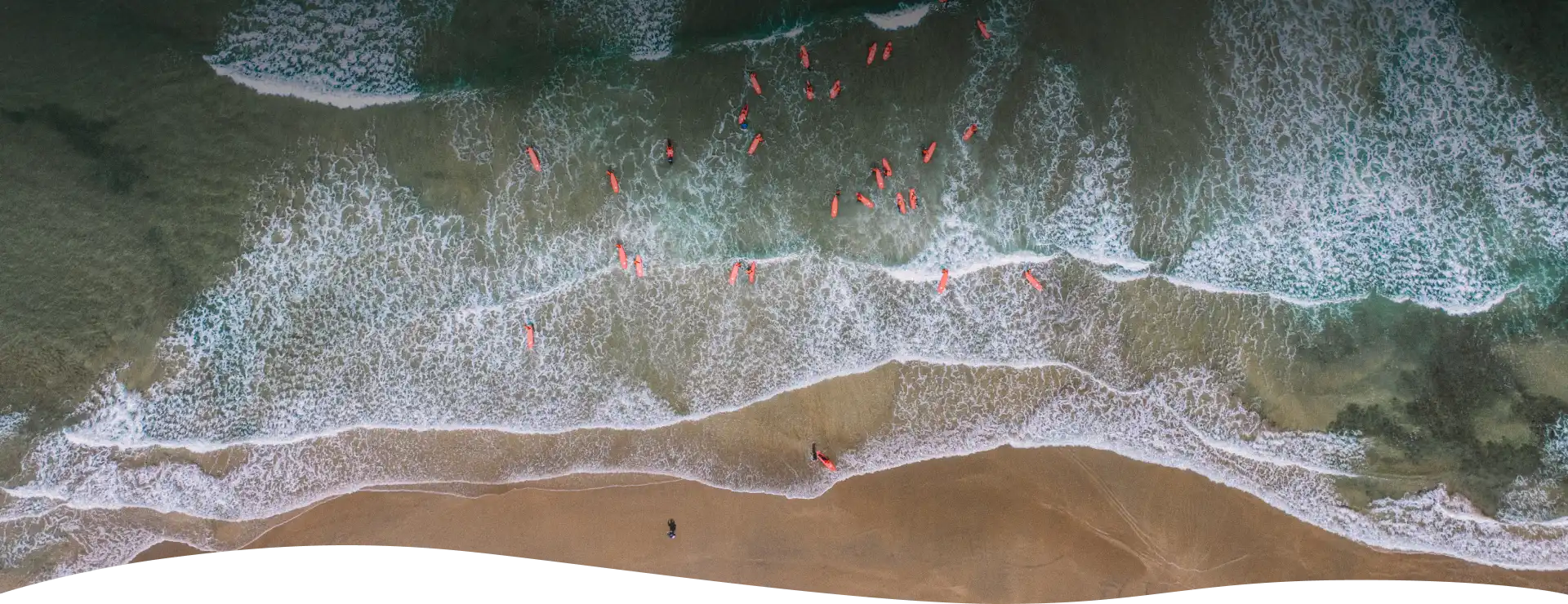 Birds view of group of people standing in semi circle with their surf boards on beach with sand dune and a city view in background