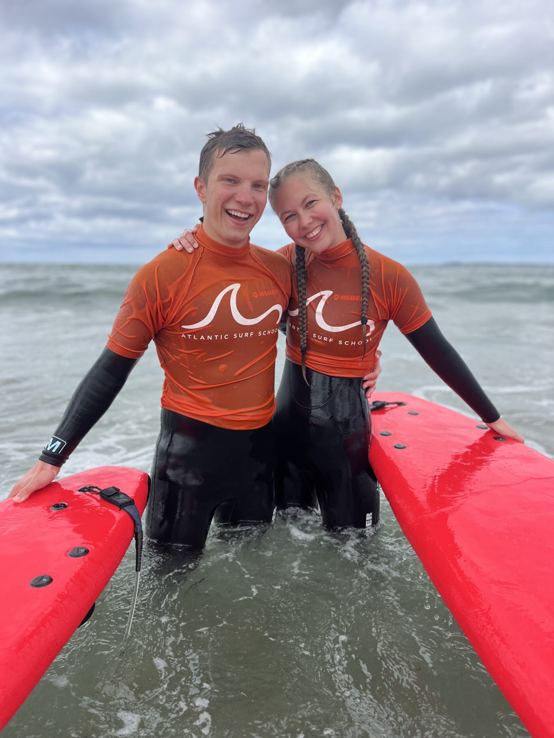Two smiling people in orange Atlantic Surf School shirts and wetsuits stand in shallow sea water, each holding a red surfboard, with waves and a cloudy sky behind them—capturing the welcoming spirit highlighted in the school's Welcome Guide.